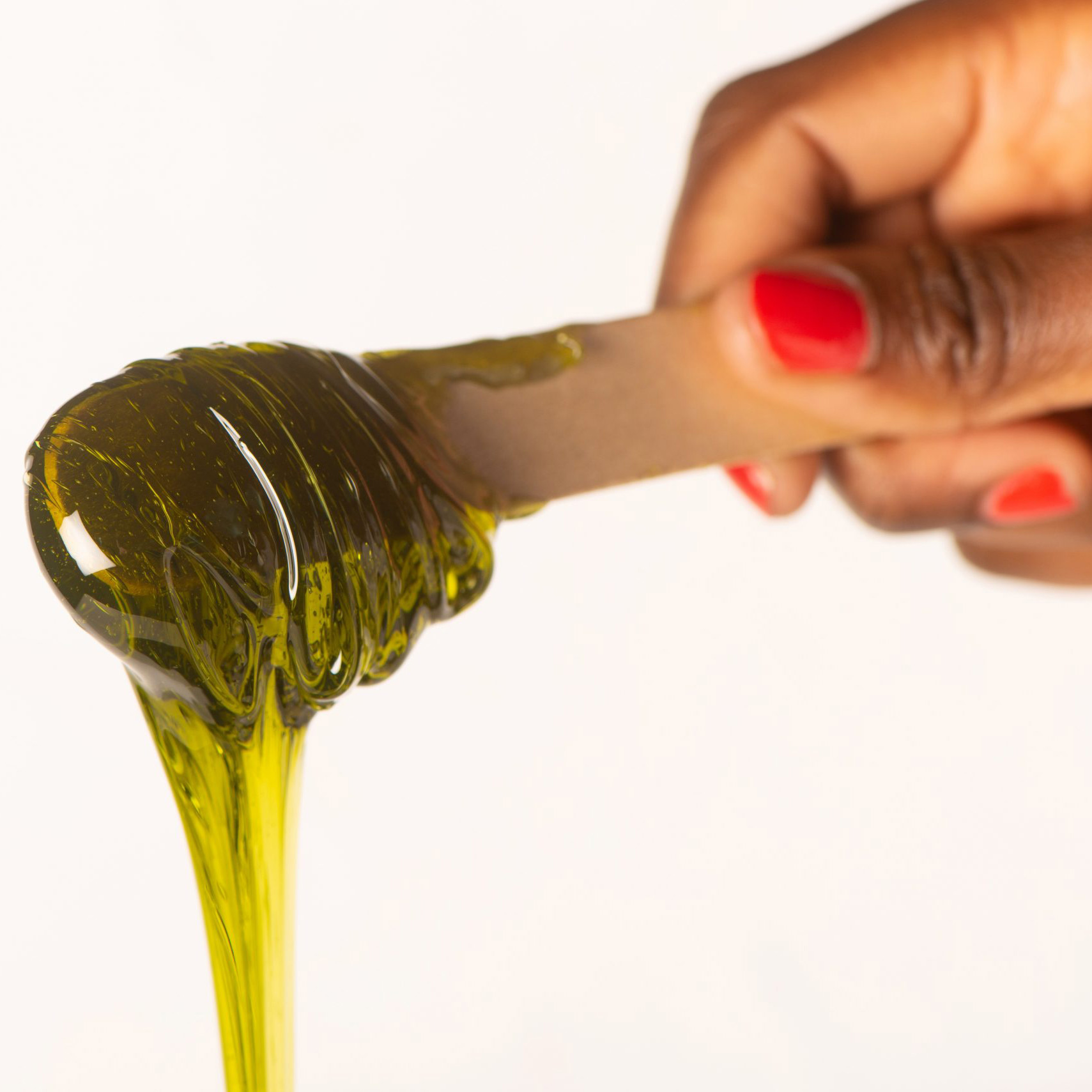 Close-up of a hand with red nails tilting a glass olive oil bottle, olive oil pouring from the spout against a white background.