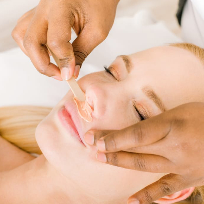 Esthetician applying a facial peel with a spatula on a woman's face, her eyes closed for treatment.