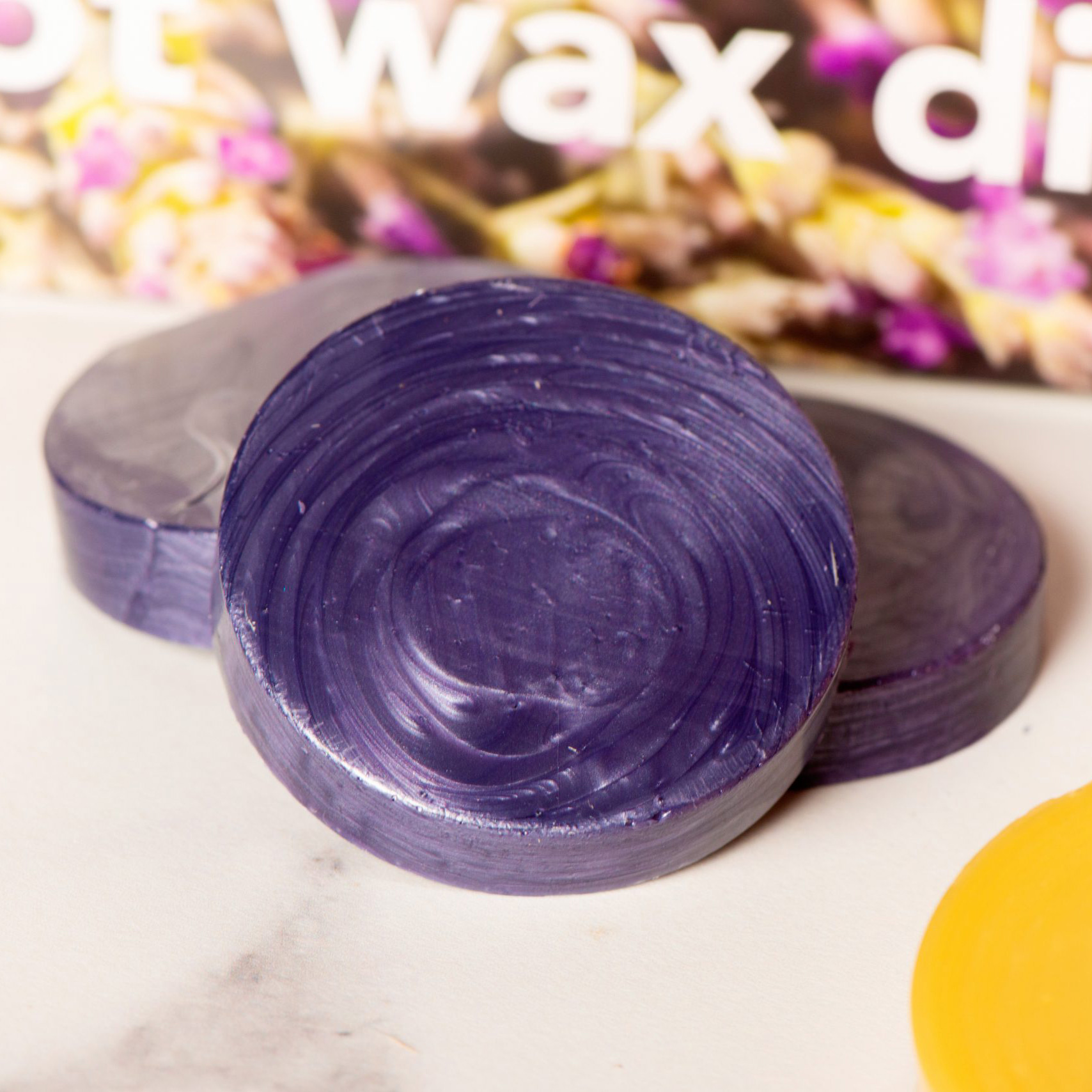Close-up of glossy purple circular soaps or discs stacked on a light surface, with a colorful blurred background behind them.