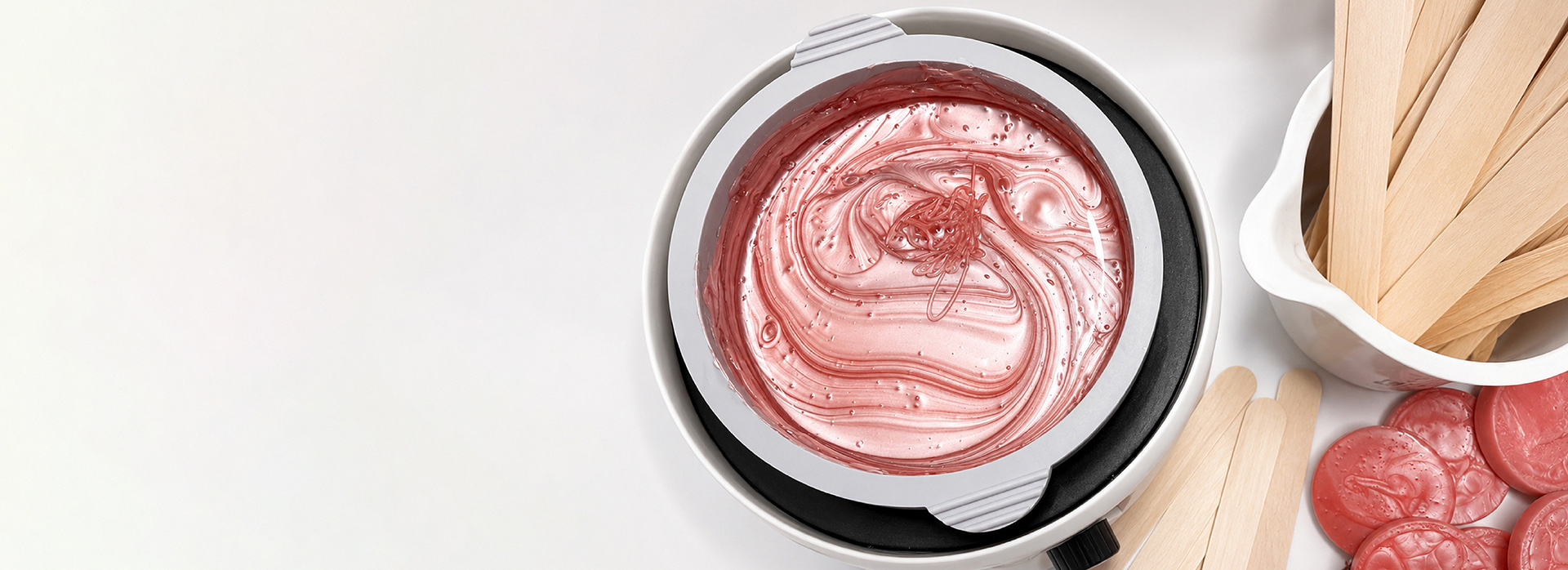 Pink melted wax in a circular warmer with a lid, surrounded by wooden spatulas and pink wax discs on a white surface.
