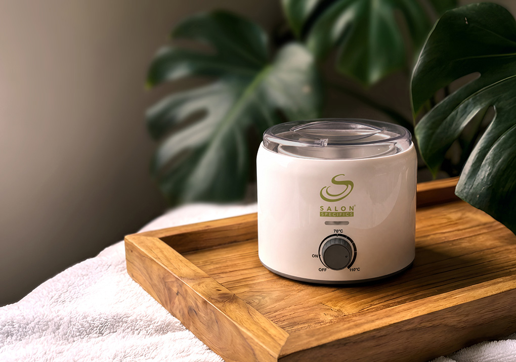 Small white wax warmer on a wooden tray in a spa-like setting with green plants in the background.