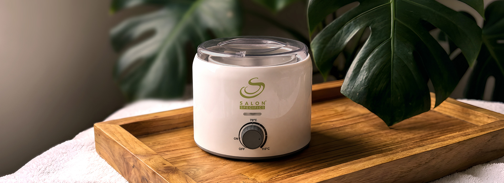 White salon wax warmer with a clear lid on a wooden tray, Salon Specifics logo visible, plant in the background.