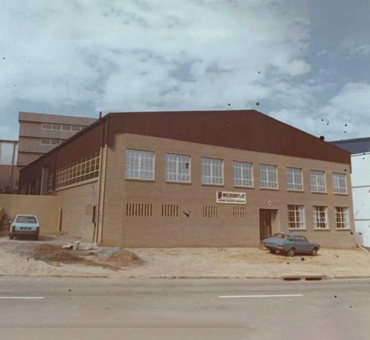 A brick industrial building with two stories, rows of large windows, a dark brown gabled roof, and a sign on the front facade, with vintage cars parked along a sandy lot in front of it under a cloudy sky.