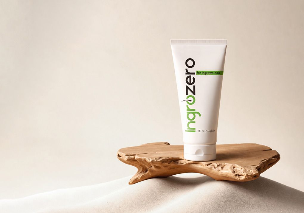 Tube labeled "ingrowZero" for ingrown hair stands on a wooden platform atop a sandy surface in a neutral studio setting, showcasing the product.