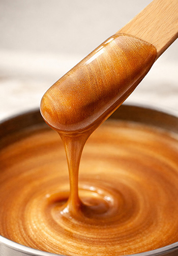 Close-up of a wooden spatula lifting thick caramel, droplets curling back into a glossy caramel pool.