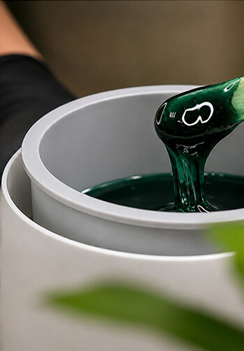 Close-up of a green glass bottle pouring viscous emerald liquid into a white cylindrical container with layered rims.
